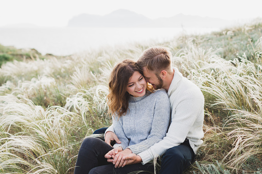 Feliz joven amante pareja sentada en el Prado de hierba de la pluma, riendo y abrazando, estilo casual suéter y jeans photo