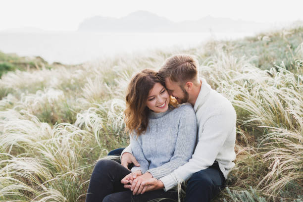 Happy young loving couple sitting in feather grass meadow, laughing and hugging, casual style sweater and jeans stock photo