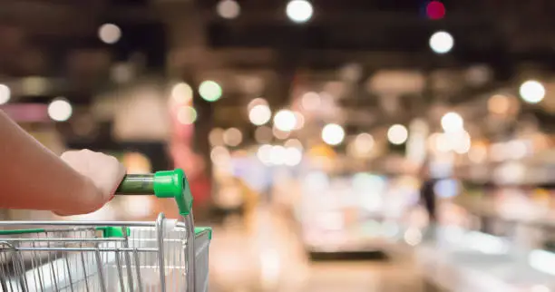 Photo of woman hand hold supermarket shopping cart with Abstract grocery store shelves blurred defocused background with bokeh light