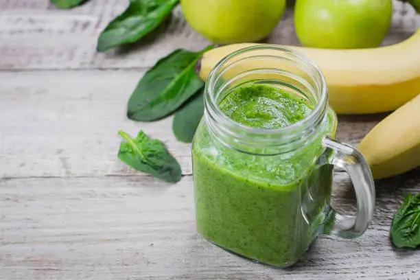 Organic Healhty Vegetarian Food. Fresh homemade raw vegetable and fruit green smoothie in mason jar with apple, banana and spinach leaves on shabby wooden background. Copy space