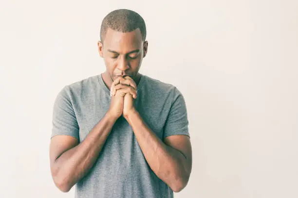 Calm spiritual handsome African guy praying with closed eyes. Serious peaceful young man with joining hands meditating. Belief concept