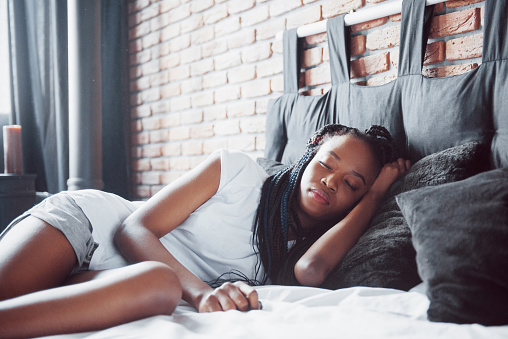 A beautiful African woman lies and sleeps in bed, in a white bedroom. Good morning, vacation at home. The girl is wearing a T-shirt, pigtails on her head.