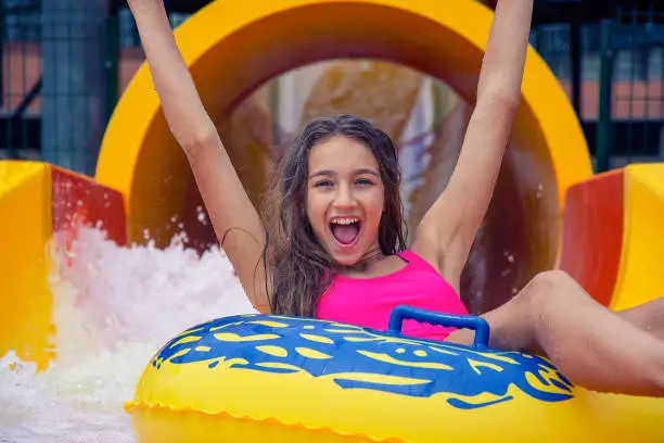 Smiling girl have fun sliding down on yellow inflatable ring with hands up in aqua park summer time concept