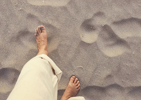 One middle aged female takes a walk on the beach.  She is barefoot and the sand is soft.  Wearing cream linen rolled up trousers.  The image taken using a mobile device from a selfie point of view.  High angle view looking down at bare feet.  Location is Ko Lanta, Krabi province, Thailand.
