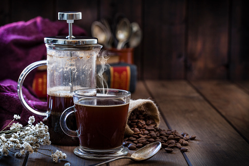 Front view of a transparent glass coffee cup and a coffee french press placed at the left of a rustic wooden table leaving useful copy space for text and/or logo at the center-right. A burlap sack with roasted coffee beans and old books at background complete the composition. Predominant color is brown. Low key DSRL studio photo taken with Canon EOS 5D Mk II and Canon EF 100mm f/2.8L Macro IS USM.