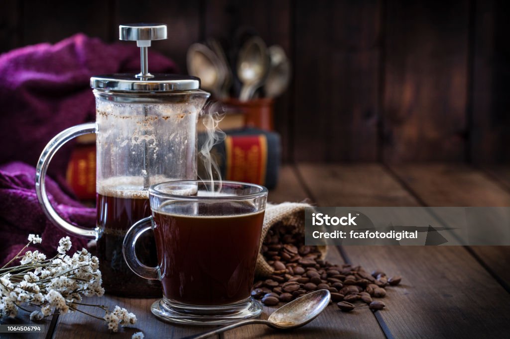 Coffee backgrounds: coffee cup and coffee french press on rustic wooden table with copy space Front view of a transparent glass coffee cup and a coffee french press placed at the left of a rustic wooden table leaving useful copy space for text and/or logo at the center-right. A burlap sack with roasted coffee beans and old books at background complete the composition. Predominant color is brown. Low key DSRL studio photo taken with Canon EOS 5D Mk II and Canon EF 100mm f/2.8L Macro IS USM. French Press Stock Photo