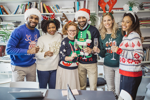 Multi ethnic group of coworkers celebrate New Year in office. They are in office holding champagne glasses, celebrating ugly sweater day.