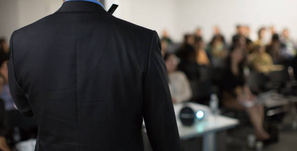 "Speaker on Stage Presenting at Event. Audience at Conference Hall Lecture Series. Corporate Presenter Presentation on Healthcare Initiatives. Crowd of People Blurred De-focused. Expert Public Speech Expert Public Speech" stock photo