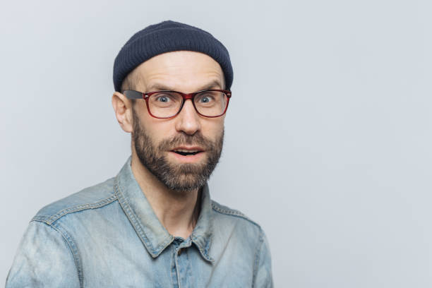 istock Stylish middle aged male with suprised and curious expression wears spectacles, denim jacket and hat, poses against grey background with copy space for your advertisment or promotional text. 1064585816