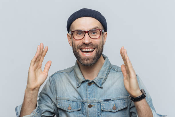 istock Handsome happy man raises hands with excitment, has overjoyed expression, looks with delighted face, has thick beard and mustache. Glad man in eyewear poses against white background, has fun 1064585920
