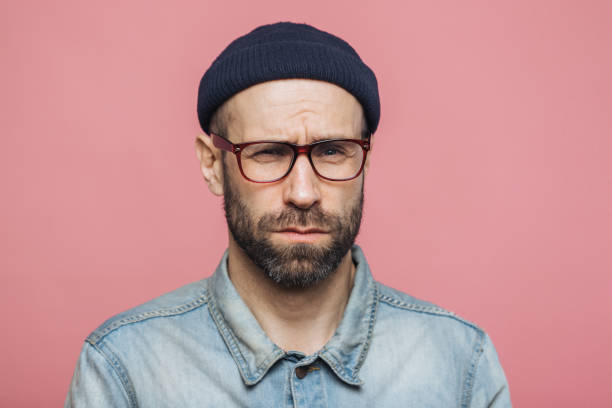 istock Unhappy discontent unshaven male looks with grumpy expression into camera, wears glasses black hat and stylish denim shirt, isolated over pink background. Displeased middle aged man poses indoor 1064585944