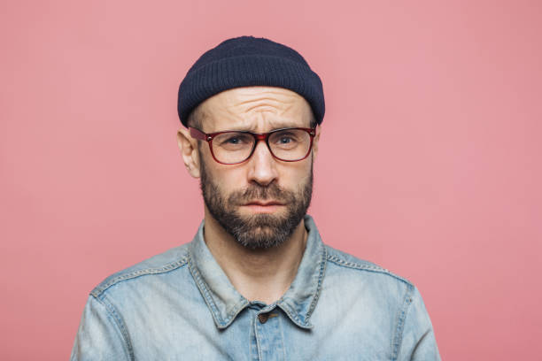 istock Grumpy bearded man with offended expression, being dissatisfied with something, frowns face, wears spectacles, hat and shirt, isolated over pink background. Displeasure and negativity concept 1064585962