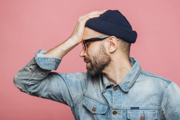 istock Sideways portrait of bearded male has regretful expression, keeps hand on forehead, looks desperately, isolated over pink background. Stressful unshaven man in glasses has terrible headache. 1064586014