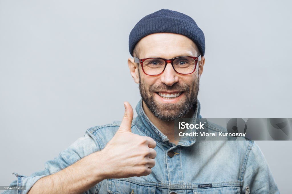 Portrait of glad middle aged male witth thick beard and mustache shows his satisfaction with something, raises thumb, has positive smile on face, isolated over white background. Happiness concept Achievement Stock Photo