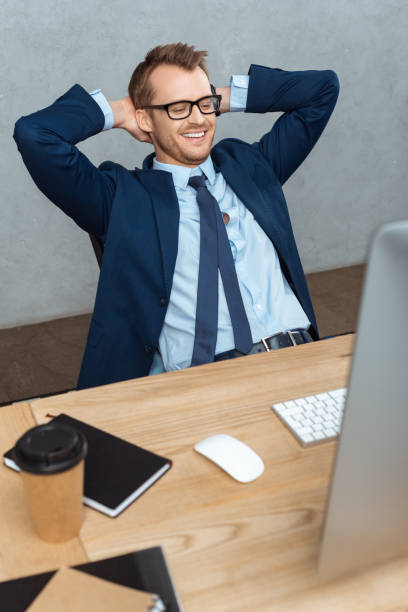istock happy young businessman in eyeglasses sitting with hands behind head at table with computer monitor in office 1064589066