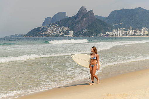 Brazilian woman in her 20's strolling by water's edge, wearing bikini, holding surf board, views of Two Brothers Mountain in background