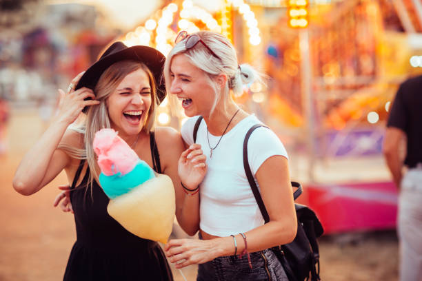 Friends in amusement park Shot of happy female friends in amusement park eating cotton candy. Two young women enjoying a day at amusement park. traveling carnival photos stock pictures, royalty-free photos & images