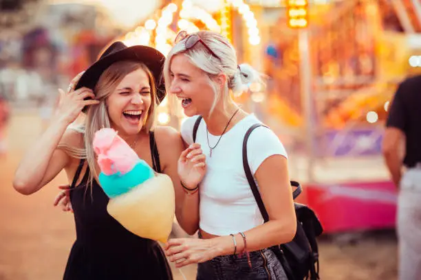 Shot of happy female friends in amusement park eating cotton candy. Two young women enjoying a day at amusement park.