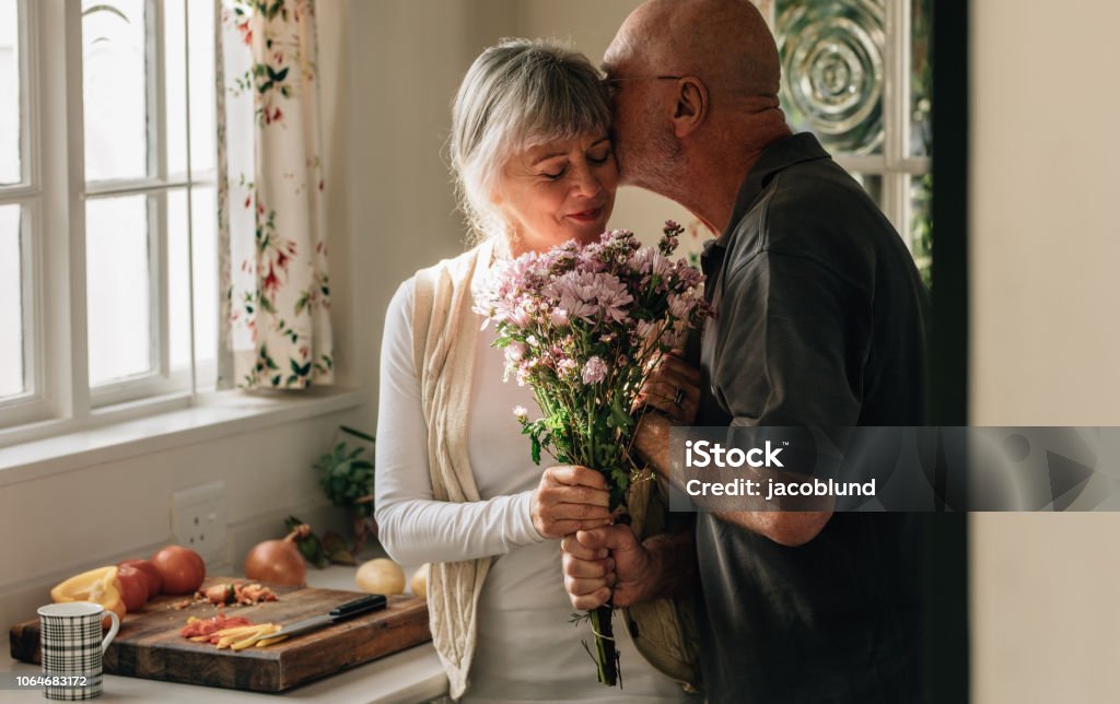 Romantic senior couple at home expressing their love Senior couple standing in kitchen holding a bunch of flowers. Senior man kissing his wife holding her hand at home. Senior Adult Stock Photo