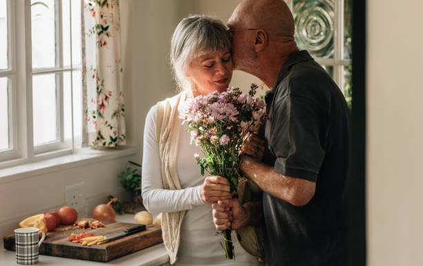 Romantic senior couple at home expressing their love Senior couple standing in kitchen holding a bunch of flowers. Senior man kissing his wife holding her hand at home. pictures of husband and wife pictures stock pictures, royalty-free photos & images