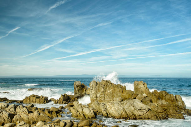 Powerful Pacific Ocean wave crashing on the rocks along the California coast, with clouds and jet contrails in the background sky.\n\nTaken in Pacific Grove, California, USA.