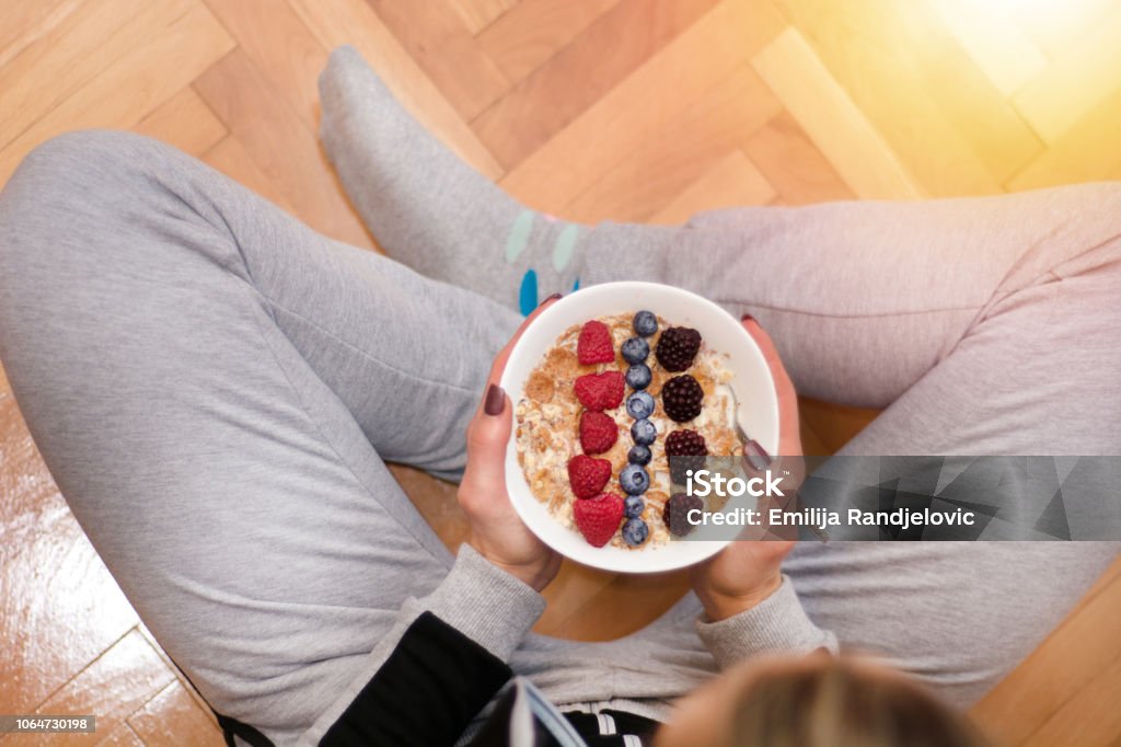 Femme en vêtements maison manger la bouillie d’avoine dans un bol orné de mûres, framboises et bleuets - Photo de Céréales du petit déjeuner libre de droits