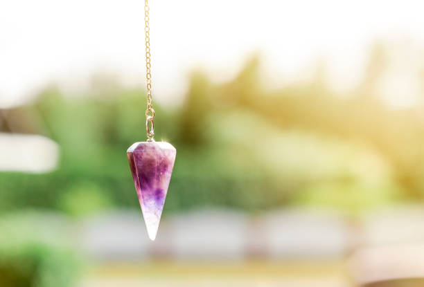 Amethyst crystal pendulum. Semi precious stone quartz pendulum on silver metal chain hanging outdoors in sunny day, green background. pendulum stock pictures, royalty-free photos & images