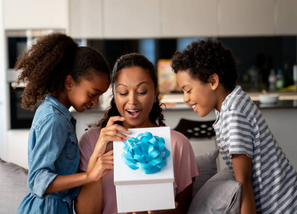 Happy kids surprising their mother with a gift for Mother's Day Happy African American kids surprising their mother with a gift at home and smiling - Mother's Day concepts anniversary photos stock pictures, royalty-free photos & images