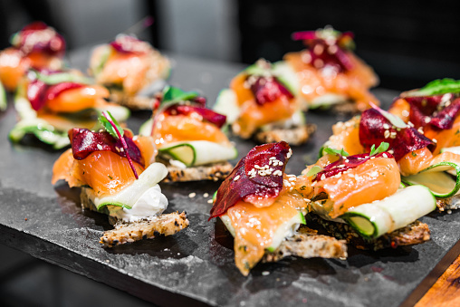 Close up of a tapa with bread, smoked salmon, zucchini, cream cheese, red beetroot, sesame seeds, and greens at a street food market fair festival