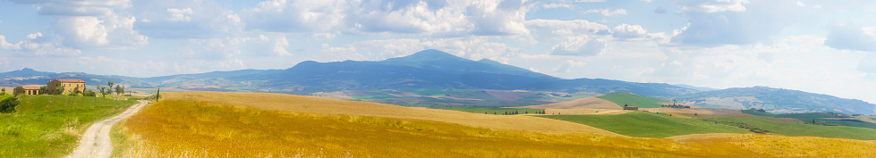 Panoramic view of Tuscany landscape, with hills, cypress trees by roads, agriculture fields, mostly with wheat, some mountains and dramatic cloudy sky in background.