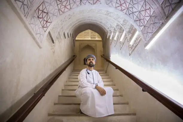 arab man in traditional omani outfit resting at the stairs of an old castle