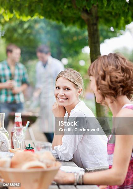 Young Couple Sitting By Picnic Table With People In The Background Stock Photo - Download Image Now