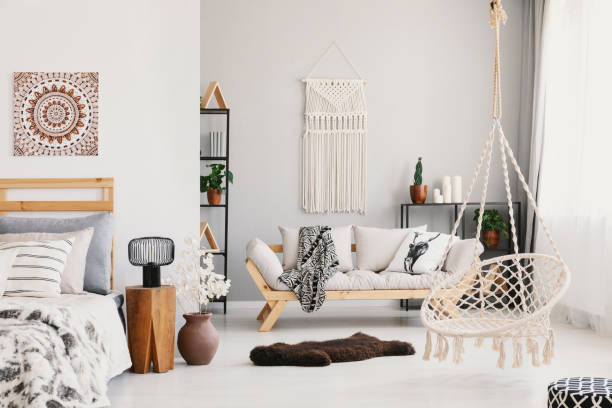 istock Bright living room interior with macrame on the wall, beige couch with pillow and blanket, hammock chair, fluffy rug and bedside table with lamp standing by the bed in the real photo 1064926202