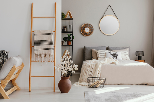 Scandi open space bedroom interior with double bed with knit blanket and many pillows, rack with books and decor, carpet on the floor in real photo