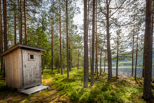 Outdoor wooden toilet in a beautiful sunny forest wilderness landscape by a lake. Horizontal composition.