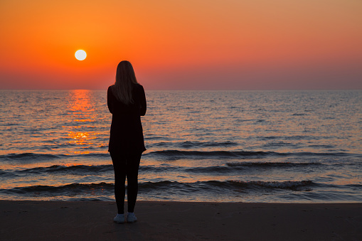 Young adult woman standing alone on sand and staring at small waves of sea and orange sunset. Girl silhouette on beach. Peaceful atmosphere in summer evening. Back view.