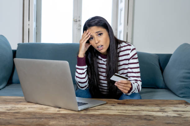Attractive young latin woman desperate and furious with credit card and laptop siting on sofa home in credit card problems debts online payment online shopping online banking bad financial situation. stock photo