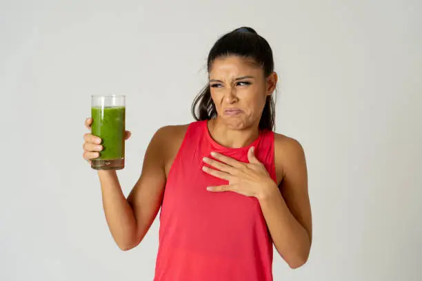 Attractive dieting girl holding detox green juice with angry negative face showing dislike gestures not wanting healthy drink isolated on grey background in healthy food rejection weight loss concept.