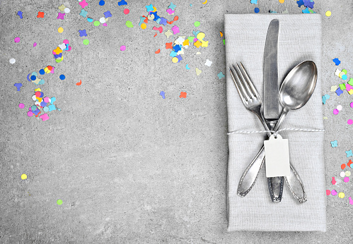 Birthday table setting background with copy space. Concrete background with napkin, silverware and name tag. Cutlery with fork, knife and spoon. Top view, party or event decoration with confetti.