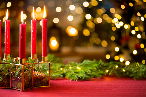 Background of glass and gold candleholder with red candles and spruce branches on red table cloth. Living room decorated with lights and candles and Christmas tree