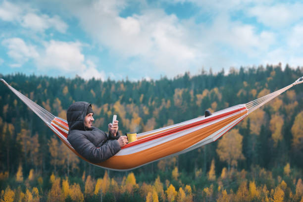 ein lächelnder mann sitzt in einer hängematte und eine smartphone in malerischer lage. becher in seiner rechten hand. - telephone nature mobile phone autumn stock-fotos und bilder