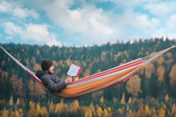 A man sits in a hammock and reads a book in a picturesque place. Mug in his right hand. A man sits in a hammock and reads a book in a picturesque place. Mug in his right hand finnish lapland autumn stock pictures, royalty-free photos & images