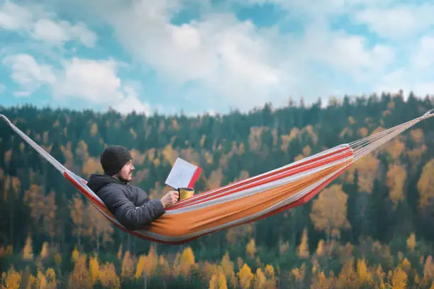 Photo of A man sits in a hammock and reads a book in a picturesque place. Mug in his right hand.