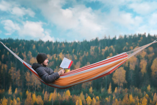 a man sits in a hammock and reads a book in a picturesque place. mug in his right hand. - forest sitting men comfortable imagens e fotografias de stock