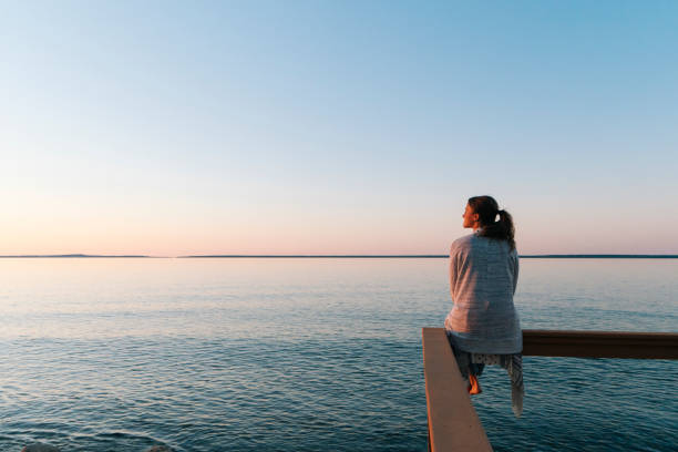 Young woman sitting on edge looks out at view sunset and sea behind, Michigan ocean life stock pictures, royalty-free photos & images