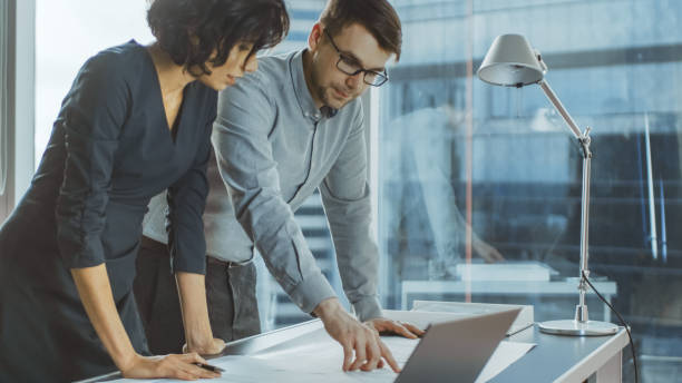 istock Two Young Business Entrepreneurs Leaning on a Desk, Working with Documents for a New Startup Project. Businesspeople Using Laptop in the Office with Cityscape Window View. 1065050000