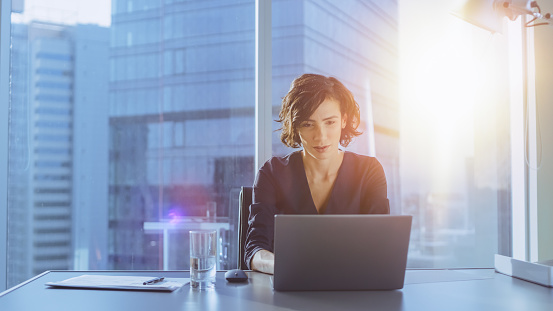 Beautiful Successful Businesswoman Working on a Laptop in Her Office with Cityscape View Window. Strong Independend Female CEO Runs Business Company. Sun Flares Behind Her.