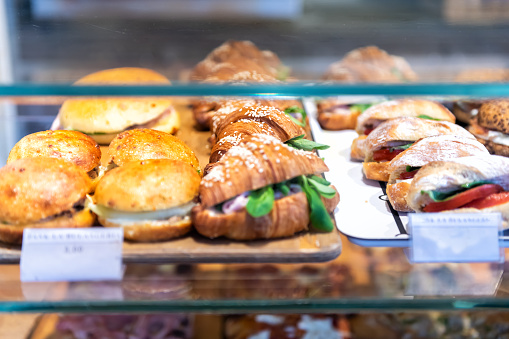 Display of store, shop selling Italian panini sandwiches with deli bologna meat, ricotta cheese, tomatoes, green spinach, buns, croissants on tray, platter