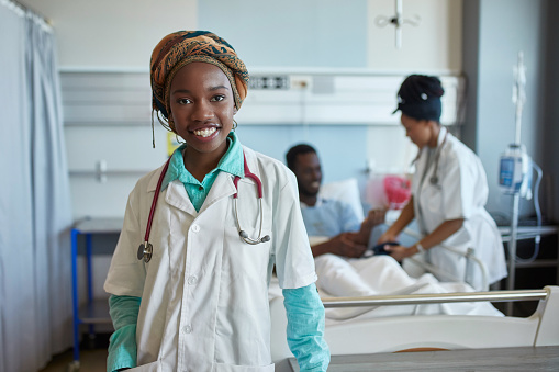 Confident female medical student with colleague examining patient in background. Portrait of smiling young trainee is in hospital ward. She is wearing lab coat.