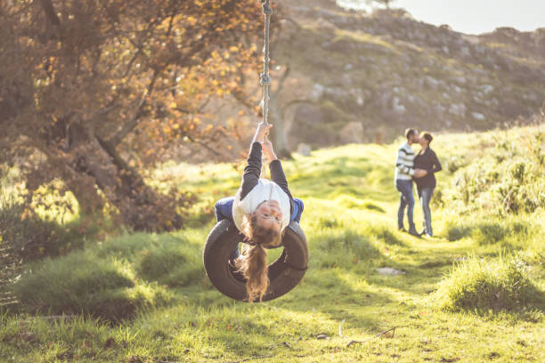 Little girl with curly brown hair, swinging on a black tyre swing with her mother and father kissing in the background, Cape Town, South Africa stock photo
